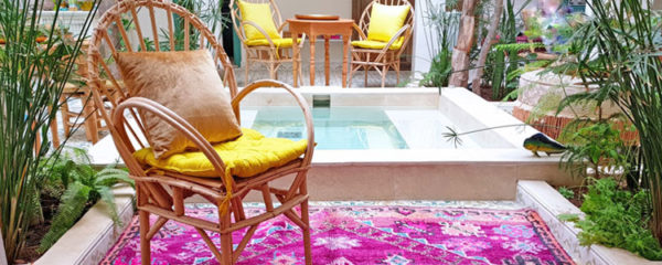 Photo d'un patio berbère en dessous d'un puits de lumière avec un tapis rose, des fauteuils en osier avec des coussins jaune et un point d'eau