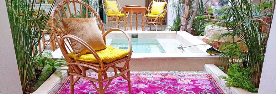 Photo d'un patio berbère en dessous d'un puits de lumière avec un tapis rose, des fauteuils en osier avec des coussins jaune et un point d'eau